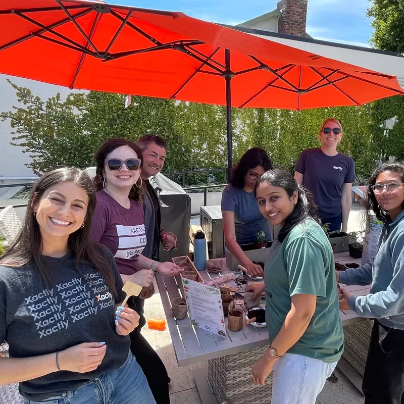 A group of diverse Xactly employees have fun in the sun under a large, red patio umbrella while completing a planting activity.