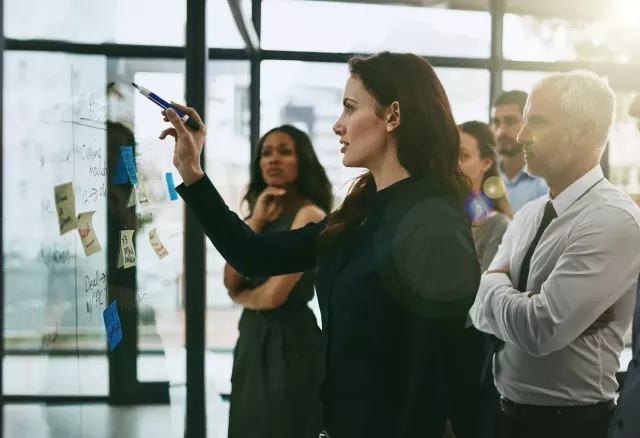 A business woman draws ideas on a glass wall as colleagues look on contemplatively.