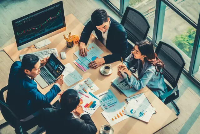 Three male and one female co-worker all in business clothing sit around a wooden table discussing charts and graphs.