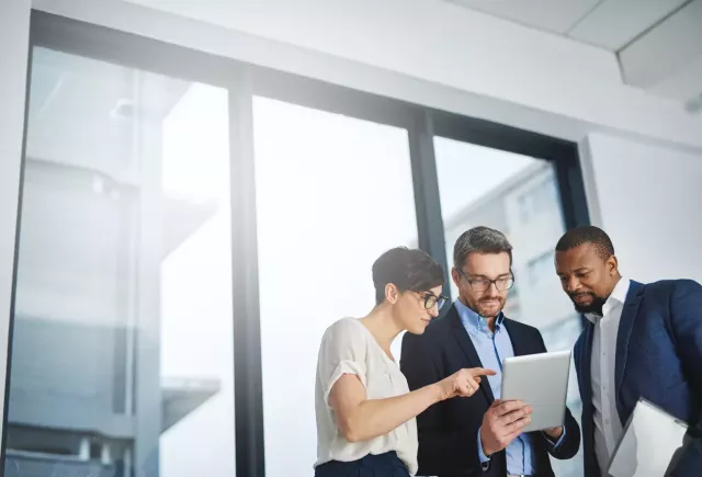 Three diverse coworkers in professional attire look at a tablet computer together while standing in front of tall transparent glass office windows with similar tall buildings visible outside in the daylight.