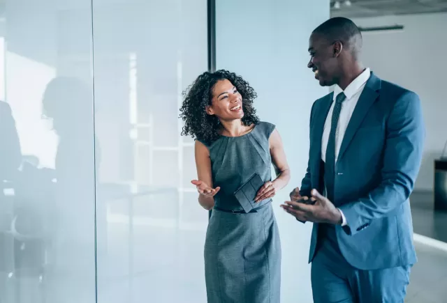 Co-workers in business attire smile and gesture in an office with the glass wall behind them reflecting a conference table and chairs.