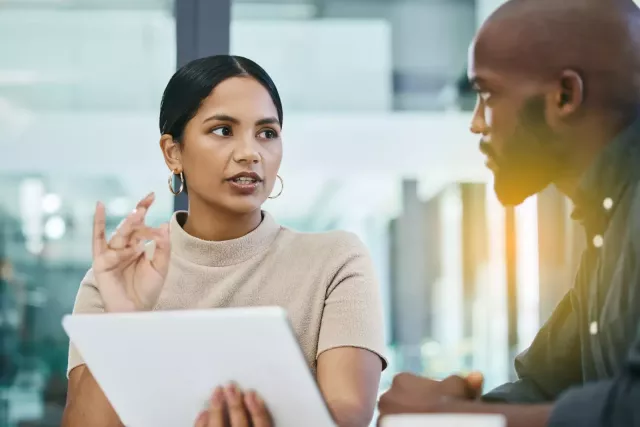 Male and female coworkers discuss sales figures at a conference table in an office with large glass windows with lens flare from sunrise/sunset in background.