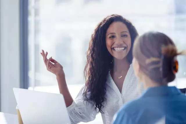 Two woman site at a table in a bright office with large glass windows. One is smiling toward the camera at the other.