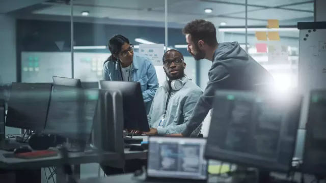 Three coders discuss software improvements in a room full of monitors displaying code.