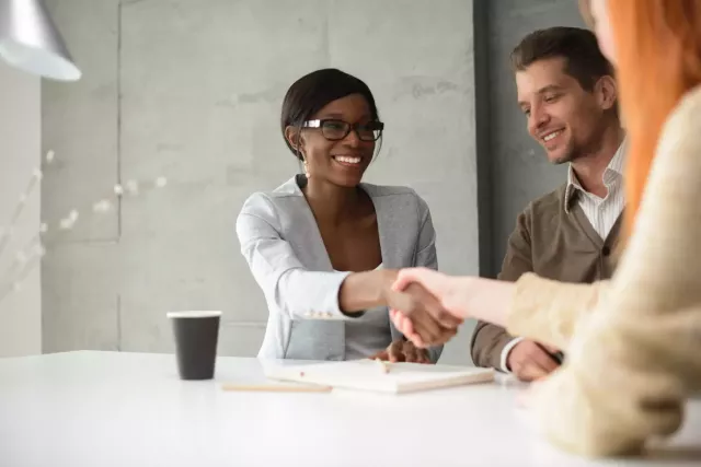 Smiling businesswoman shakes hands with female colleague  over a conference table, while male colleagues smiles in the background.