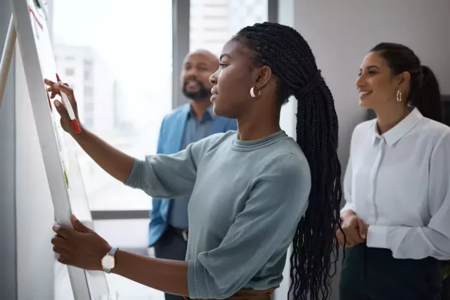 Diverse group of young professionals gathers to discuss buyer personas in a modern office with one woman working on a white board.