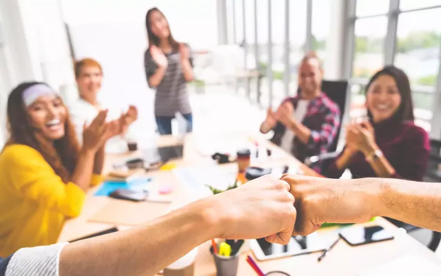 Sales team gathered around a conference table clap in congratulations while two hands fist-bump in the foreground.