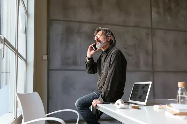 A man in a modern, industrial-style office sits on his desk and looks out the window while talking on the phone.